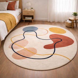 Puede incluir: Una alfombra redonda con un diseño abstracto. La alfombra presenta un fondo crema con formas coloridas en azul, amarillo, melocotón y óxido. Una línea azul oscuro atraviesa las formas. La alfombra está colocada sobre un suelo de madera.