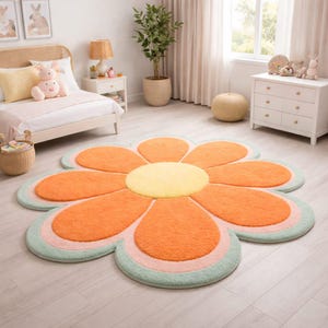 May include: A large, flower-shaped rug with orange petals, a yellow center, and a light green border. The rug is placed on a light-colored wooden floor in a bedroom setting. The room also features a bed, a dresser, and a potted plant.