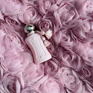 Pode incluir: Um frasco de perfume rosa claro com tampa prateada e detalhe de borla repousa sobre uma cama de rosas de tecido macio e franzido. O frasco tem um design ornamentado. O fundo é um mar de rosas de tecido rosa.