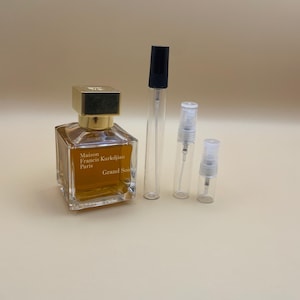 Puede incluir: Un frasco de perfume y tres viales de muestra. El frasco de perfume es cuadrado con una tapa dorada y contiene un líquido dorado. El texto "Maison Francis Kurkdjian Paris Grand Soir" está impreso en el frasco.