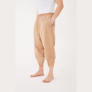 May include: Tan-colored cropped pants with an elastic waistband and pockets. The pants have a unique design with a split hem at the ankle. The person is also wearing a white tank top. The pants are made of a soft, flowing fabric.