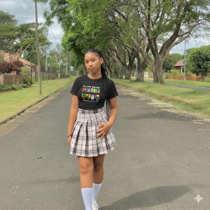 Puede incluir: Una persona con una camiseta negra con el texto "ELITE SQUAD" y un diseño gráfico, combinada con una falda plisada a cuadros. El atuendo se completa con calcetines blancos y zapatillas deportivas. El fondo presenta una calle arbolada.