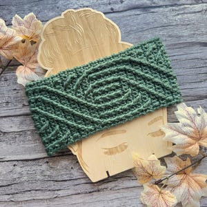 May include: A sage green crocheted headband with a textured cable knit design. The headband is displayed on a wooden cutout of a face, with fall leaves around it. The headband is approximately 4 inches wide.
