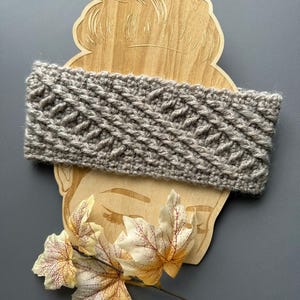 May include: A gray knitted headband with a textured pattern. The headband is displayed on a wooden cutout of a face, with decorative fall leaves in the foreground. The headband is approximately 4 inches wide.