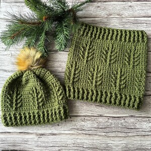Puede incluir: Un gorro verde tejido a ganchillo con un pompón de piel sintética y un cuello a juego. El gorro y el cuello presentan un patrón de hojas texturizadas. El conjunto se exhibe sobre una superficie de madera con una ramita de hoja perenne.