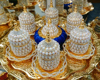 May include: Ornate gold-colored serving set with multiple cups and saucers. Each cup and lid is encrusted with faux pearls and rhinestones. The set includes a blue velvet base and is displayed on a decorative gold tray.