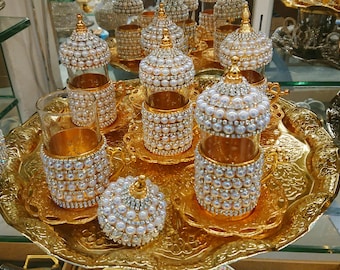 May include: A set of ornate, gold-colored tea cups and saucers, embellished with faux pearls. The set includes several cups, saucers, and lids, all arranged on a decorative gold tray. The cups have a clear glass interior.