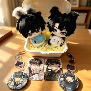 Puede incluir: Dos muñecos de peluche con cabello negro y orejas de animal dentro de un recipiente blanco con relleno de papel amarillo. Incluye dos tarjetas rectangulares con ilustraciones de anime, dos chapas redondas y dos llaveros de acrílico.
