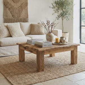 Puede incluir: Una mesa de centro cuadrada de madera rústica con acabado natural descansa sobre una alfombra de yute tejida. Un sofá blanco con cojines de colores neutros se encuentra en el fondo. Libros y un jarrón están colocados sobre la mesa, creando un ambiente acogedor.