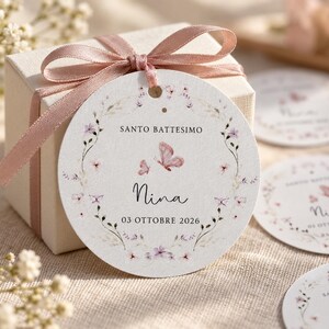 Può includere: Una scatola regalo quadrata color bianco sporco legata con un nastro di raso rosa polvere e un cartellino rotondo in carta. Il cartellino presenta una ghirlanda floreale, due farfalle rosa e il testo SANTO BATTESIMO, Nina e 03 OTTOBRE 2026.