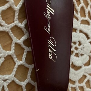 May include: A burgundy shoehorn with the gold text "Montgomery Ward" in a cursive font. The shoehorn is on a white crocheted surface.