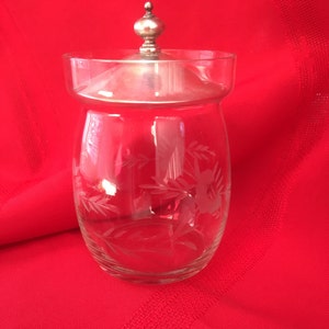 May include: A clear glass jar with a silver lid and etched floral design. The jar is on a red surface.