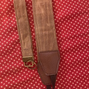 May include: A brown camera strap with a brown leather pad and a metal ring. The strap is on a red and white patterned fabric.