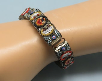 Può includere: Un braccialetto vintage realizzato con tessere di mosaico colorate. Il braccialetto è composto da piccole tessere rotonde in tonalità di rosso, blu, giallo e bianco. Le tessere sono disposte a formare un motivo di fiori e foglie. Il braccialetto è fissato con una chiusura in metallo.