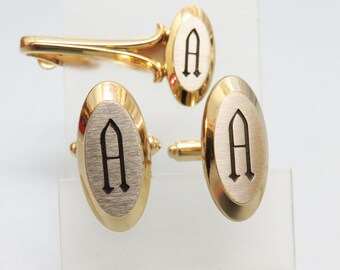 May include: A gold-toned tie clip and cufflink set. The cufflinks and tie clip have an oval design with a silver-colored center and a black letter "A". The edges of the cufflinks and tie clip are gold-colored.