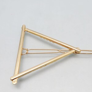 May include: A gold-colored hair barrette in a triangular shape. The barrette has a simple, geometric design with a hinged clasp for securing hair. The metal has a shiny finish, reflecting light against a neutral background.