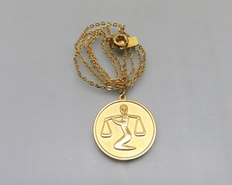 May include: A gold chain necklace with a round pendant featuring a stylized image of the astrological sign Libra.