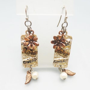 Pode incluir: Um par de brincos pendentes com acabamento prateado e cobre. Cada brinco apresenta um charme retangular com as palavras "Love Laugh" e um pequeno charme de flor. Os brincos são acentuados por uma pérola e um charme de folha.