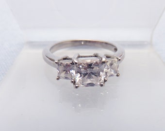 May include: A silver ring with three square-shaped clear stones. The center stone is larger than the two stones on either side.