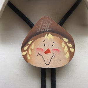 以下が含まれることがあります： 手描きの木製のかかしの顔のペンダントが付いたボロタイ。かかしは格子模様の茶色の帽子、黄色い髪、バラ色の頬、小さなオレンジ色の三角形の鼻をしており、黒い編み込みコードに取り付けられています。
