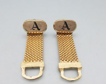 May include: A pair of gold-tone cufflinks with a chain design. Each cufflink features a black letter 'A' on a gold oval.