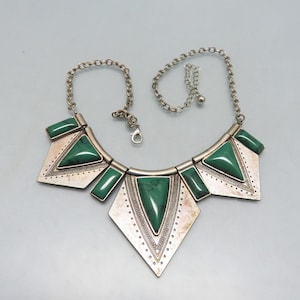 May include: Silver-toned statement necklace featuring geometric shapes and vibrant green accents. The necklace has a chain with an adjustable length and a lobster clasp. The design includes triangles, rectangles, and a diamond shape, all with a textured finish.