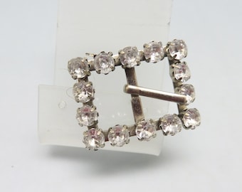 May include: A silver-toned buckle with a square frame adorned with clear rhinestones.