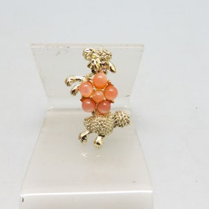 May include: A gold-toned brooch in the shape of a poodle with a cluster of pink and peach colored stones in the center.