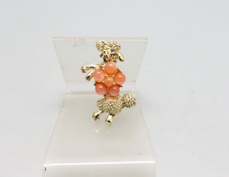 May include: A gold-toned brooch in the shape of a poodle with a cluster of pink and peach colored stones in the center.
