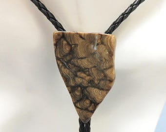 May include: A bolo tie with a triangular pendant. The pendant is made of a light brown material with a swirling, wood-grain pattern. The tie features a black braided leather cord.