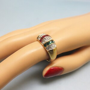 May include: Gold ring with a band of clear, red, and green stones. The stones are set in a raised, curved design.