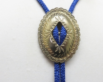 May include: A gold bolo tie with a decorative, ornate, oval-shaped pendant. The pendant has a diamond-shaped cutout in the center. The tie is made of blue cord.
