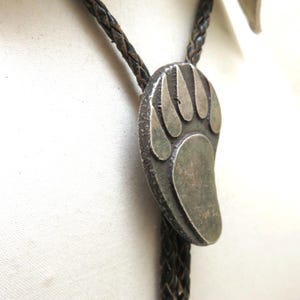 May include: A vintage bolo tie featuring a silver-toned bear paw pendant. The pendant is attached to a braided leather cord. The bear paw design has a detailed outline and is set against a textured background. The bolo tie is a classic accessory.
