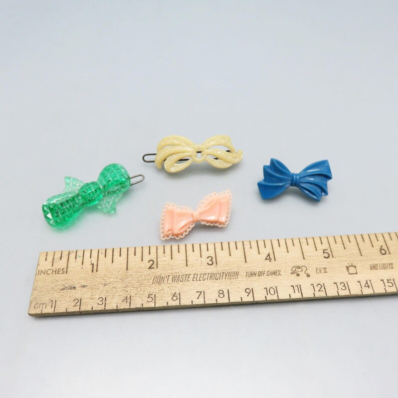 May include: A collection of vintage hair accessories, including a green clip, a yellow bow clip, a peach bow clip, and a blue bow. A wooden ruler with inch and centimeter measurements is also present. The ruler is approximately 6 inches long.