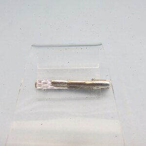 May include: Silver tie clip with a textured design. The clip is closed and ready to be attached to a tie.