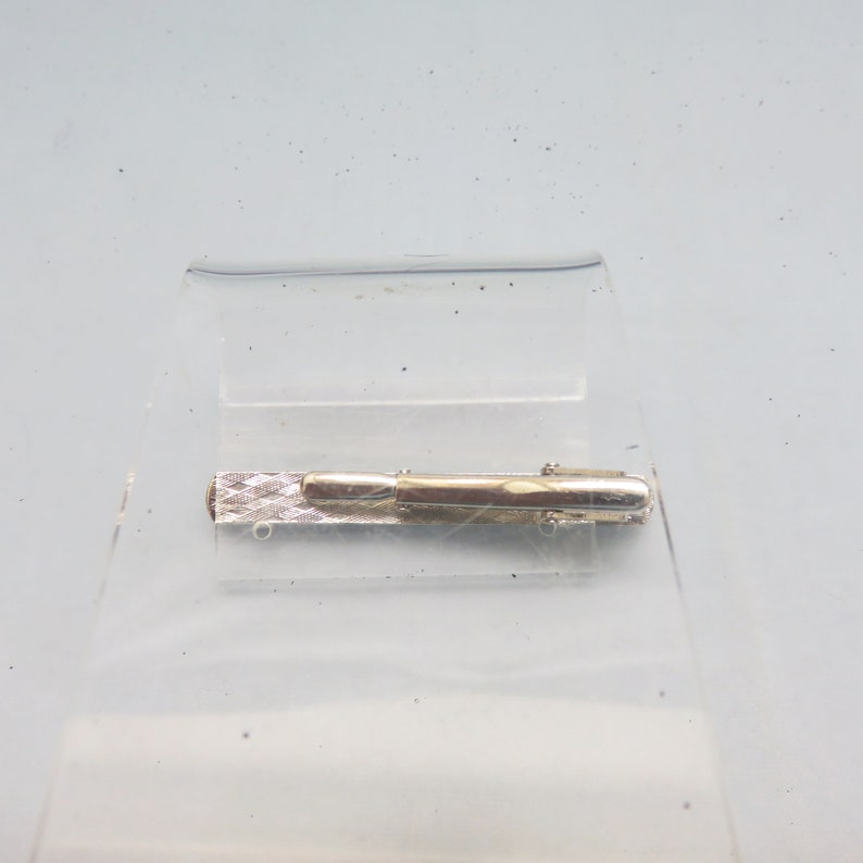 May include: Silver tie clip with a textured design. The clip is closed and ready to be attached to a tie.