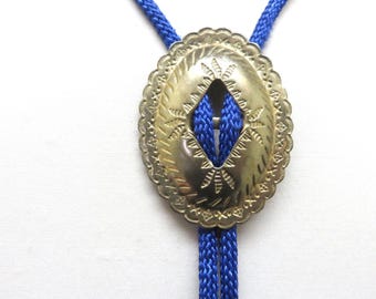 May include: A bolo tie featuring a bright blue braided cord and an oval-shaped gold-toned metal slide. The slide has a scalloped edge and is decorated with engraved geometric patterns and a central diamond-shaped cutout through which the cord passes.