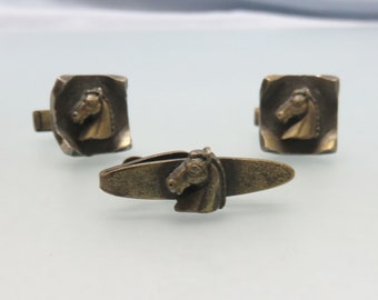 Könnte beinhalten: Ein Set aus Vintage-Manschettenknöpfen und Krawattenklammer aus Messing. Die Manschettenknöpfe sind quadratisch mit einem Pferdekopf-Design. Die Krawattenklammer hat ein Pferdekopf-Design.