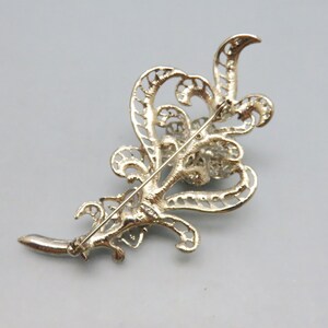May include: A silver filigree brooch with a large, round, clear rhinestone center. The brooch is in the shape of a stylized leaf.