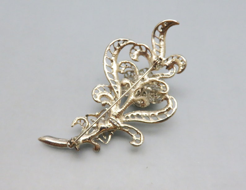 May include: A silver filigree brooch with a large, round, clear rhinestone center. The brooch is in the shape of a stylized leaf.