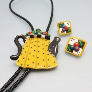 May include: A yellow teapot bolo tie with black polka dots and a black and white checkered trim. The teapot has a black spout and handle. The bolo tie comes with a pair of matching square earrings with black and white checkered trim and colorful flowers.