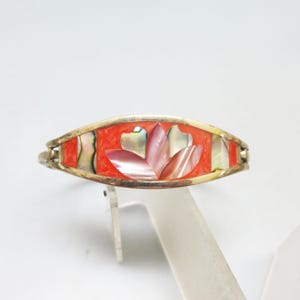 May include: A silver-toned bracelet with a central design of a pink and white flower. The flower is set against a red background, framed by iridescent shell pieces. The bracelet has a hinged clasp.