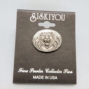 May include: A silver-colored oval pin featuring a detailed bear's head with an open mouth and sharp teeth. The pin is set against a dark gray card with the text "SISKIYOU" at the top and "Fine Pewter Collector Pins" and "MADE IN USA" at the bottom.