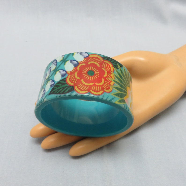 Op de afbeelding: Een turquoise bangle armband met een bloemenpatroon. Het patroon heeft een grote oranje bloem met gele en groene accenten.