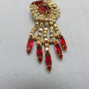 Pode incluir: Um broche dourado com um grupo de cristais vermelhos e transparentes. O broche tem um design pendente com v&aacute;rios cristais vermelhos em forma de l&aacute;grima.