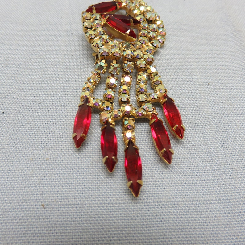Pode incluir: Um broche dourado com um grupo de cristais vermelhos e transparentes. O broche tem um design pendente com v&aacute;rios cristais vermelhos em forma de l&aacute;grima.
