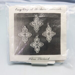 Op de afbeelding: Een zwart-witfoto van vijf zilveren sneeuwvlokversieringen. De versieringen hebben de vorm van een kruis met een decoratief ontwerp. De foto is op een witte achtergrond met de tekst "Easy/Elegant Heirloom Ornaments" en "Silver Starburst" afgedrukt op de pagina.