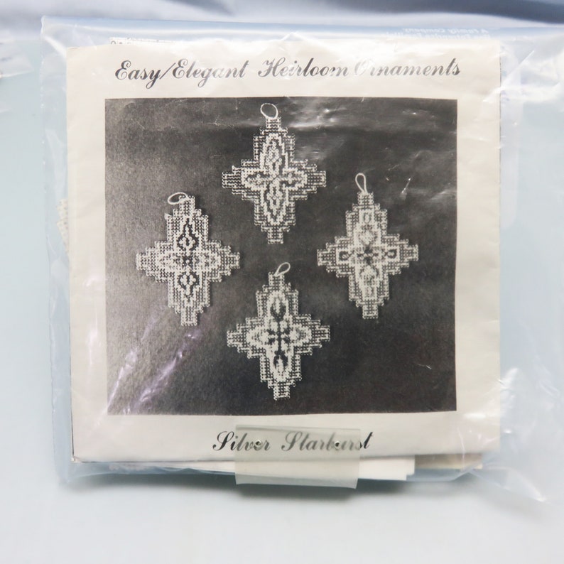 Op de afbeelding: Een zwart-witfoto van vijf zilveren sneeuwvlokversieringen. De versieringen hebben de vorm van een kruis met een decoratief ontwerp. De foto is op een witte achtergrond met de tekst "Easy/Elegant Heirloom Ornaments" en "Silver Starburst" afgedrukt op de pagina.