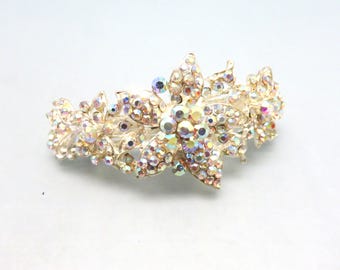 May include: A gold-toned hair clip featuring a floral design. The clip is adorned with numerous iridescent rhinestones, creating a sparkling effect. The floral elements are arranged in a symmetrical pattern, suitable for formal occasions.