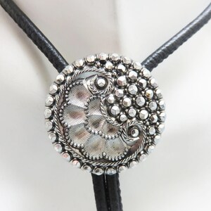May include: A silver bolo tie with a detailed, circular pendant. The pendant features a textured design with a beaded border and a black leather cord. The bolo tie is a classic accessory.
